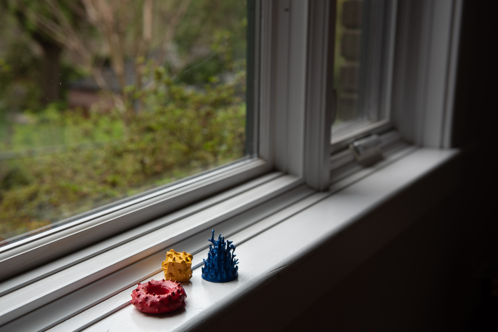Full View - three small shapes sit on a windowsill: a spiky red donut, a yellow cube with wiggly protrusions, and a blue cone with tree-like branches.
